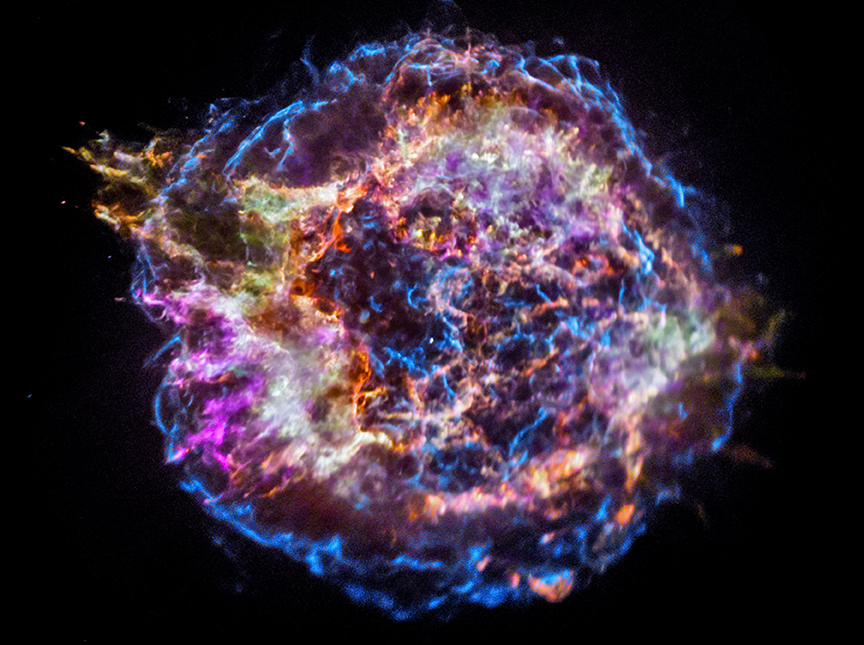 This X-ray image of Cassiopeia A resembles a disk of electric blue light, purple clouds, glowing white fog, and red and yellow flames, dotted with glowing orange specks. This is Cassiopeia A, a supernova remnant. Here, elements of the exploded star are being cast into space. The red and yellow flames are silicon and sulfur. The light purple within the cloud is iron, and the blast wave is blue. All were observed by Chandra's X-ray Observatory.